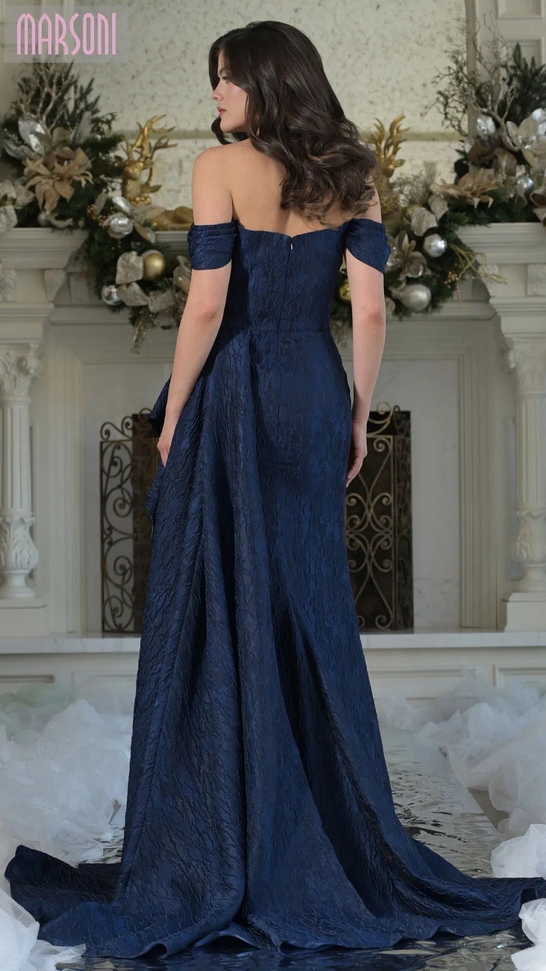 A woman in the Marsoni MV1365 long formal dress stands with her back to the camera before a decorated fireplace, the elegantly draped navy off-the-shoulder gown creating a timeless look.